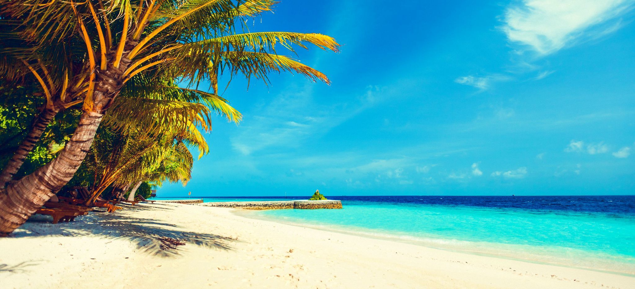 Ein Malediven-Strand zwischen türkisem Meer und Palmen. In der Ferne ein paar Wasservillen.