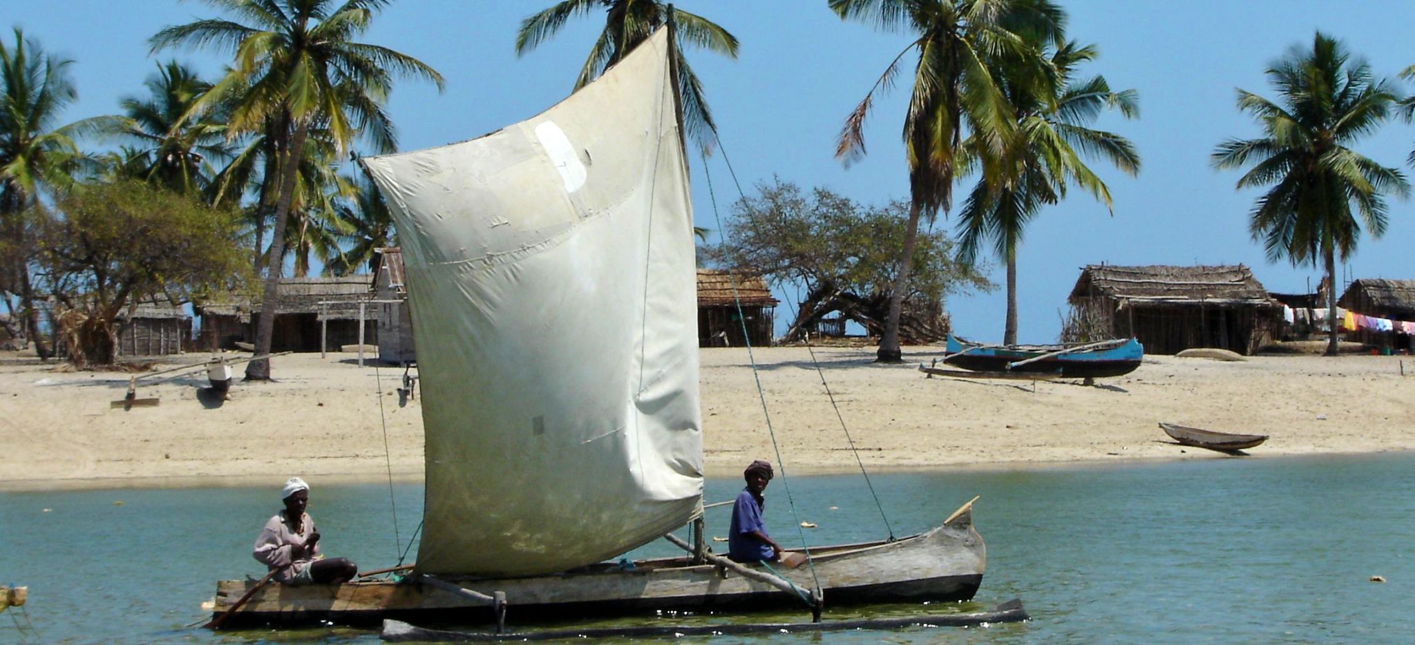 Zwei Männer auf einem traditionellen Segelboot vor Madagaskar fahren an einem Strand vorbei