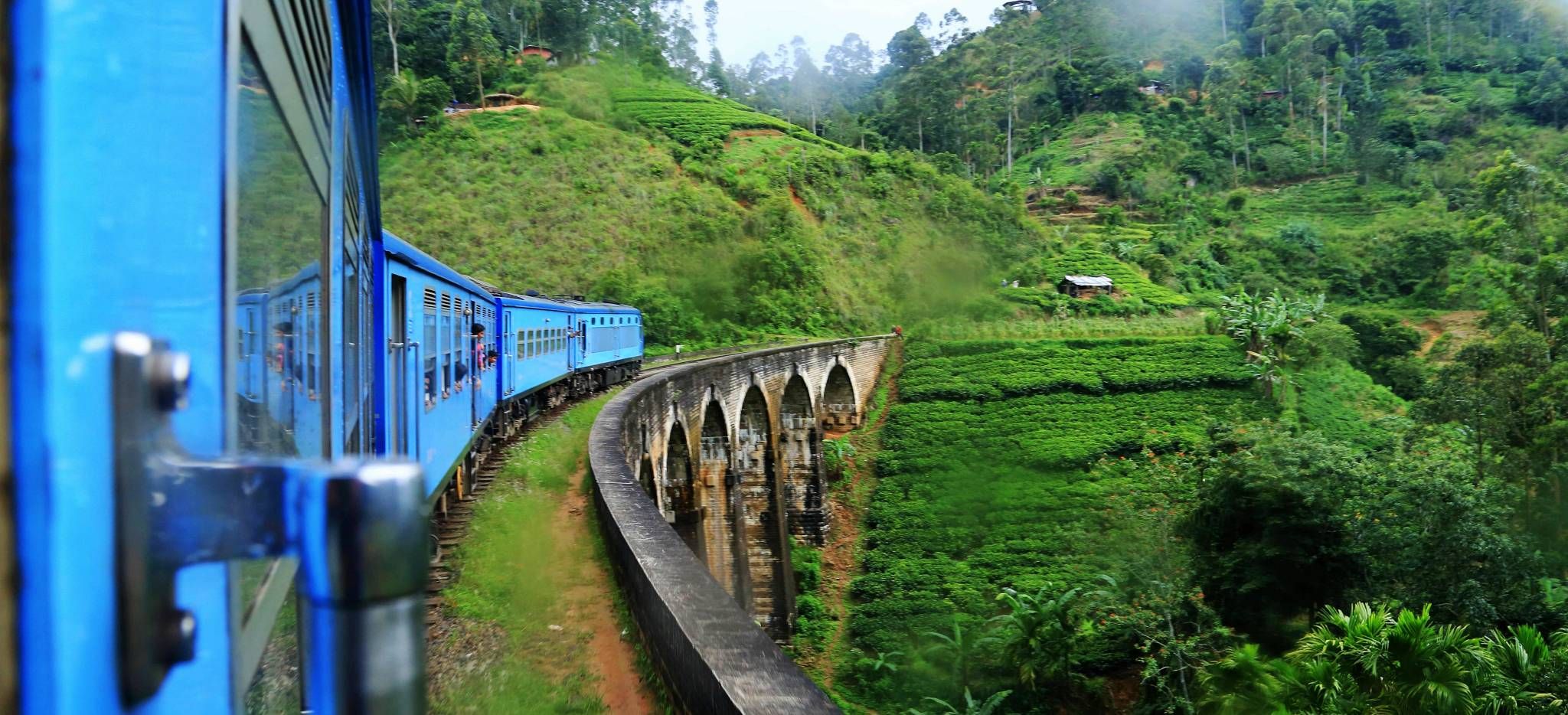 Ein blauer Zug fährt im Regen um die Kurve über eine Brücke in den Bergen von Sri Lanka 