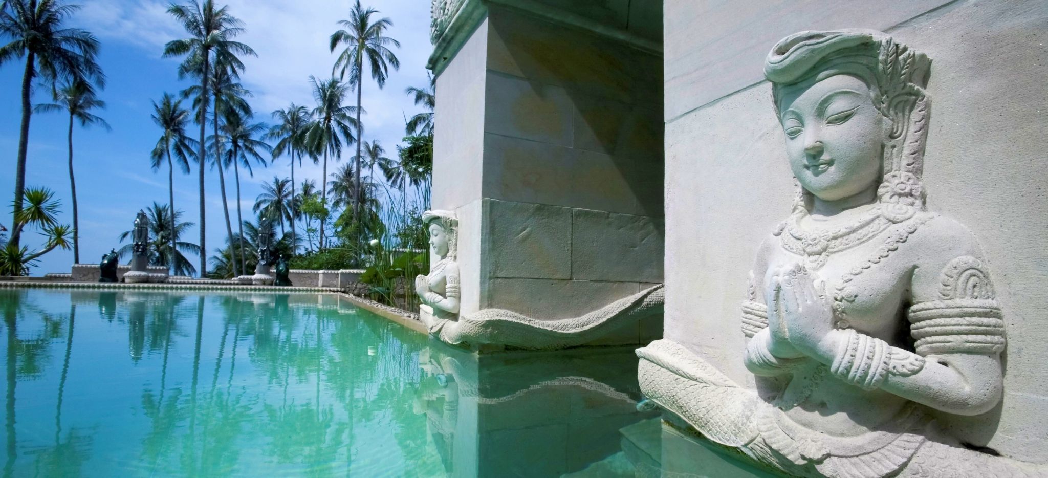 Buddhistische Statue in den Stein um einen Pool geschlagen, im Hintergrund Palmen. Das Hotel Kamalya auf Koh Samui in Thailand