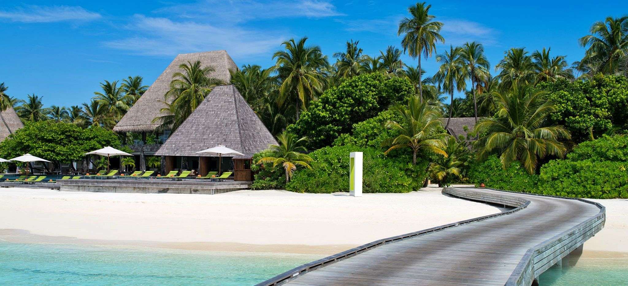 Ein Steg an einer Malediveninsel, Blick auf den Strand und mit Palmblättern gedeckte Häuser. Das Hotel Anantara Kihavah