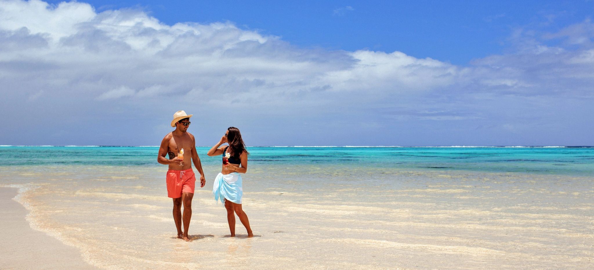 Paar am Strand Titikaveka Beach des Little Polynesian Resorts mit Cocktails in der Hand vor dem 