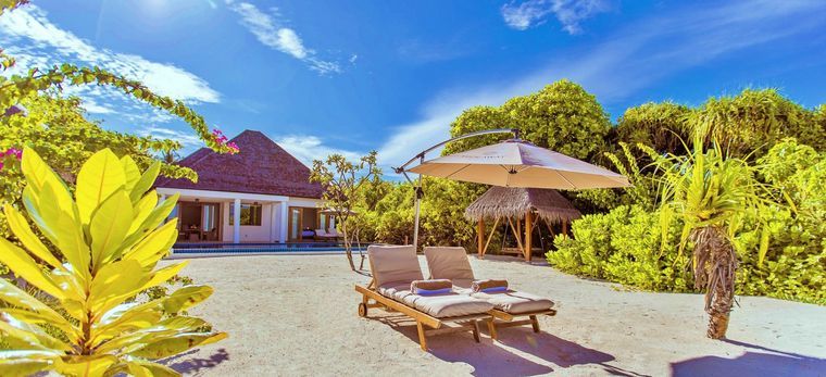 Ein Bungalow im Gebüsch mit Strand davor, auf dem sich Strandliegen befinden